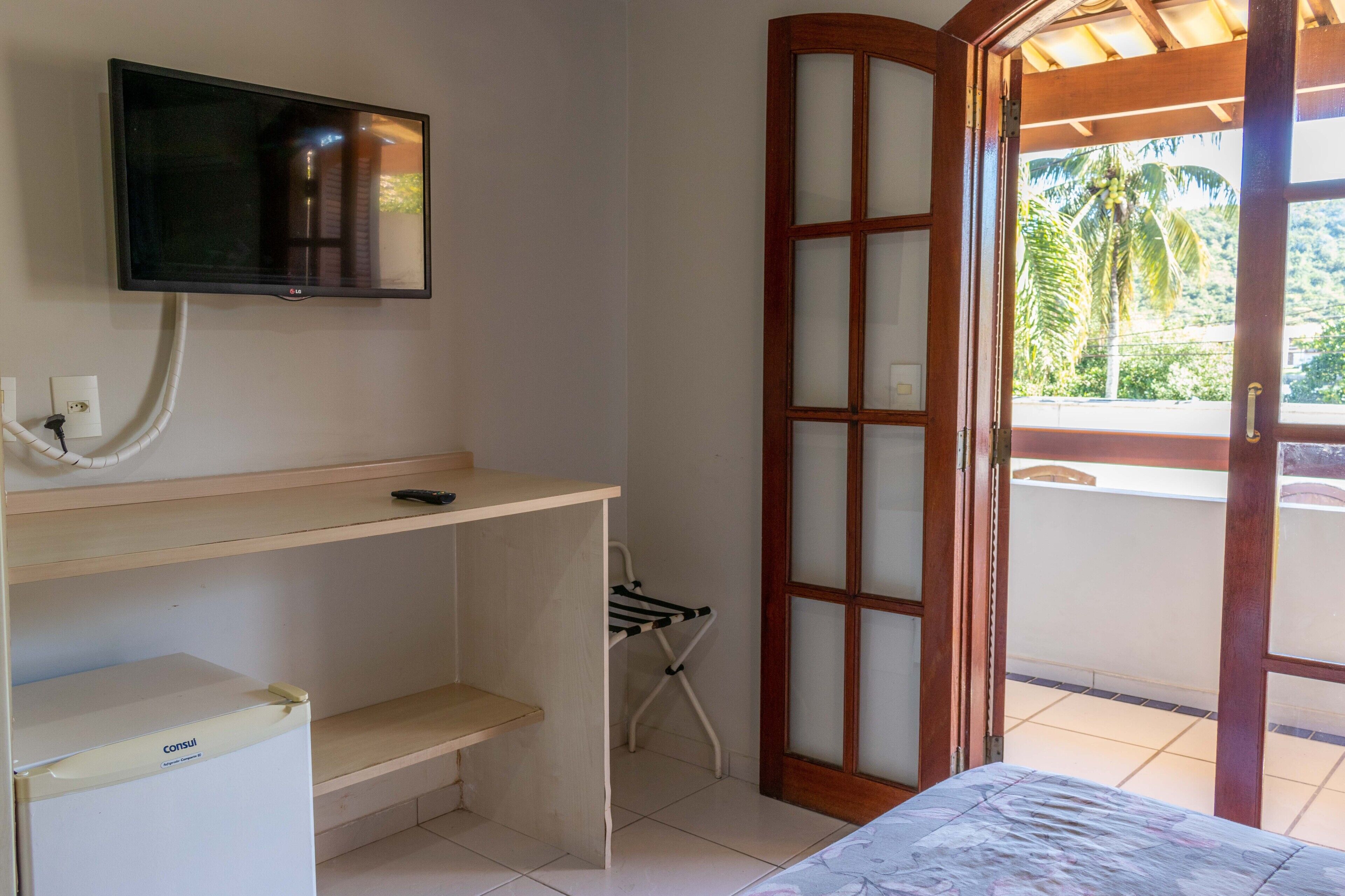 Habitación con televisión montada en la pared, un escritorio pequeño, refrigerador, una silla plegable y una puerta corrediza de vidrio que da a un balcón con vistas a palmeras.