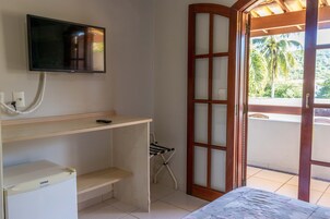 Habitación con televisión montada en la pared, un escritorio pequeño, refrigerador, una silla plegable y una puerta corrediza de vidrio que da a un balcón con vistas a palmeras.