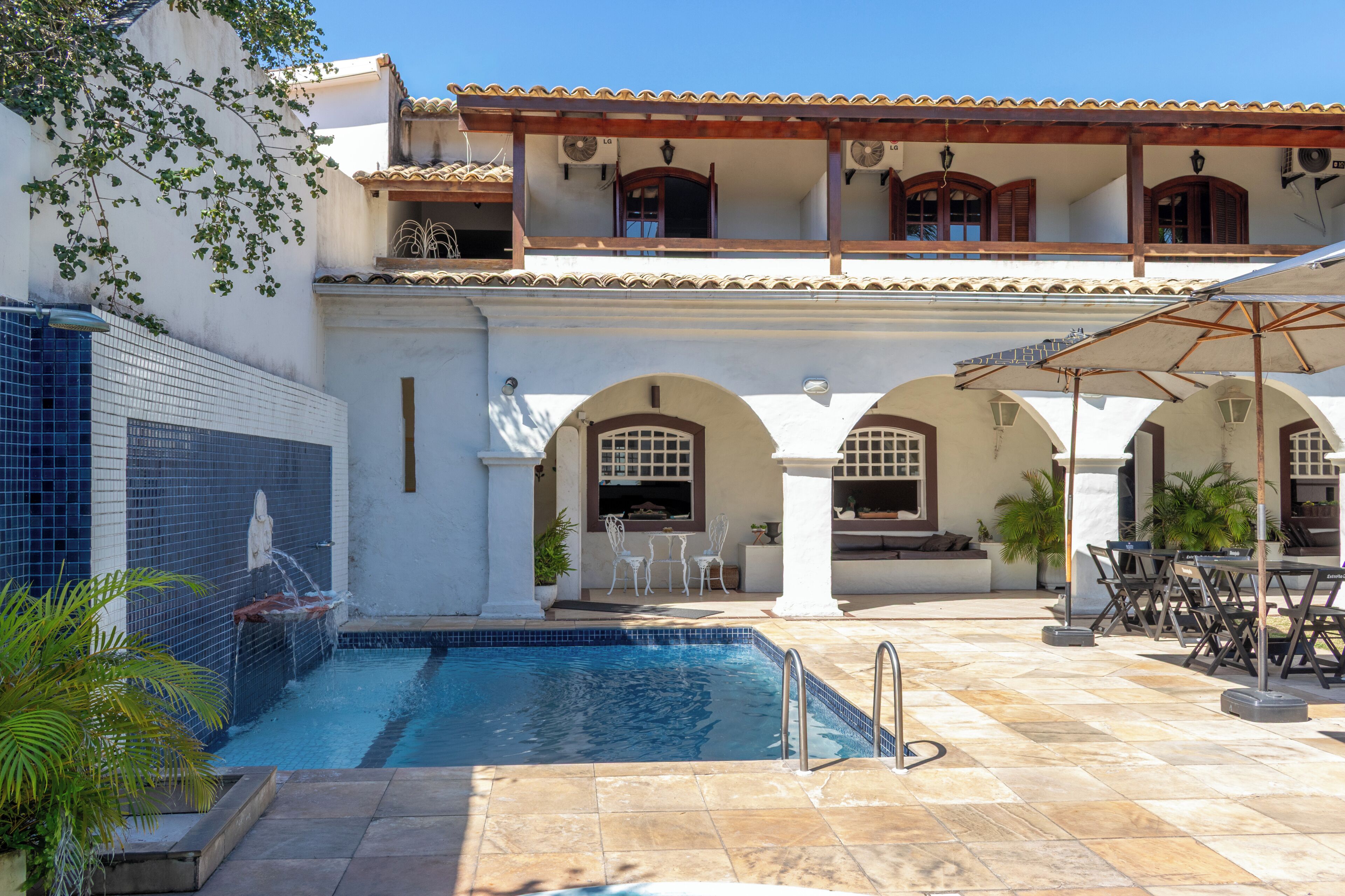 Un área de piscina con suelo de baldosas, fuente de agua y mobiliario de exterior.