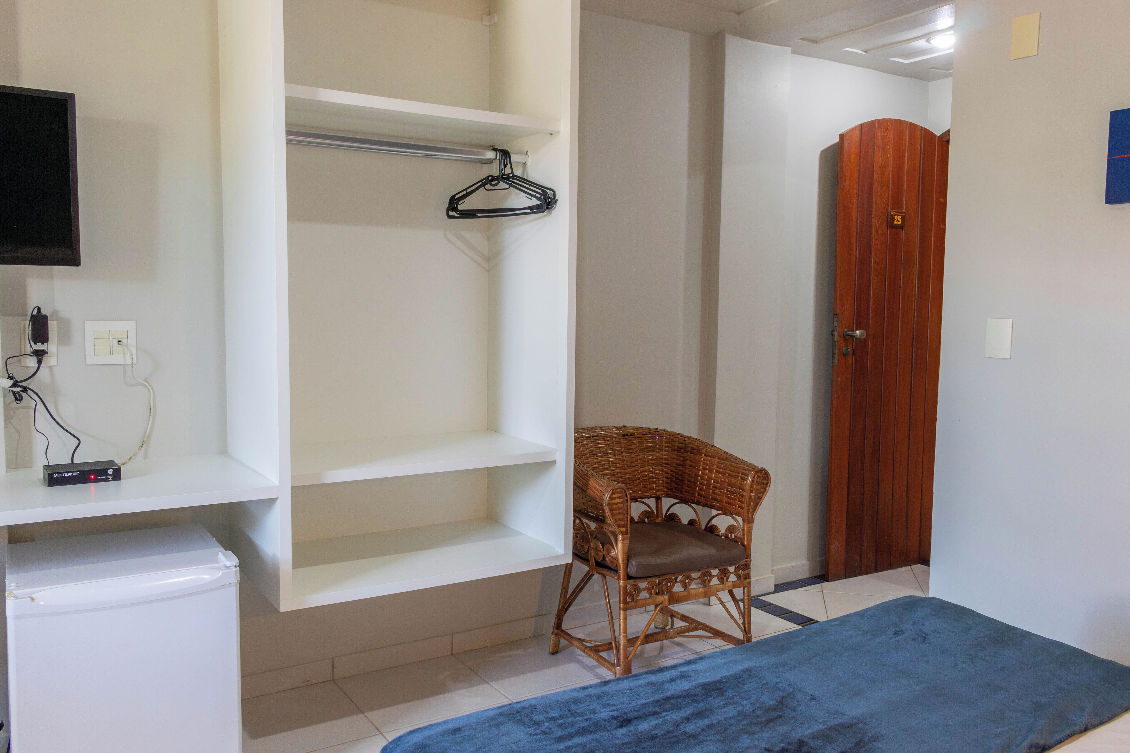 A room with a white wardrobe, a wicker chair with a cushion, a blue bedspread, and a television mounted on the wall.