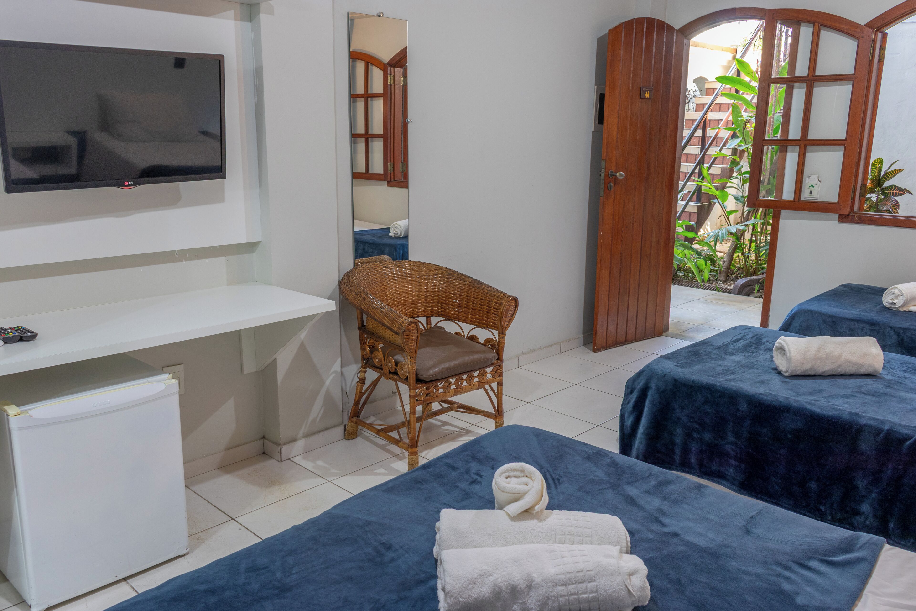 Habitación de hotel con televisión, refrigerador pequeño, silla de mimbre, dos camas con toallas y una puerta que da a un balcón.