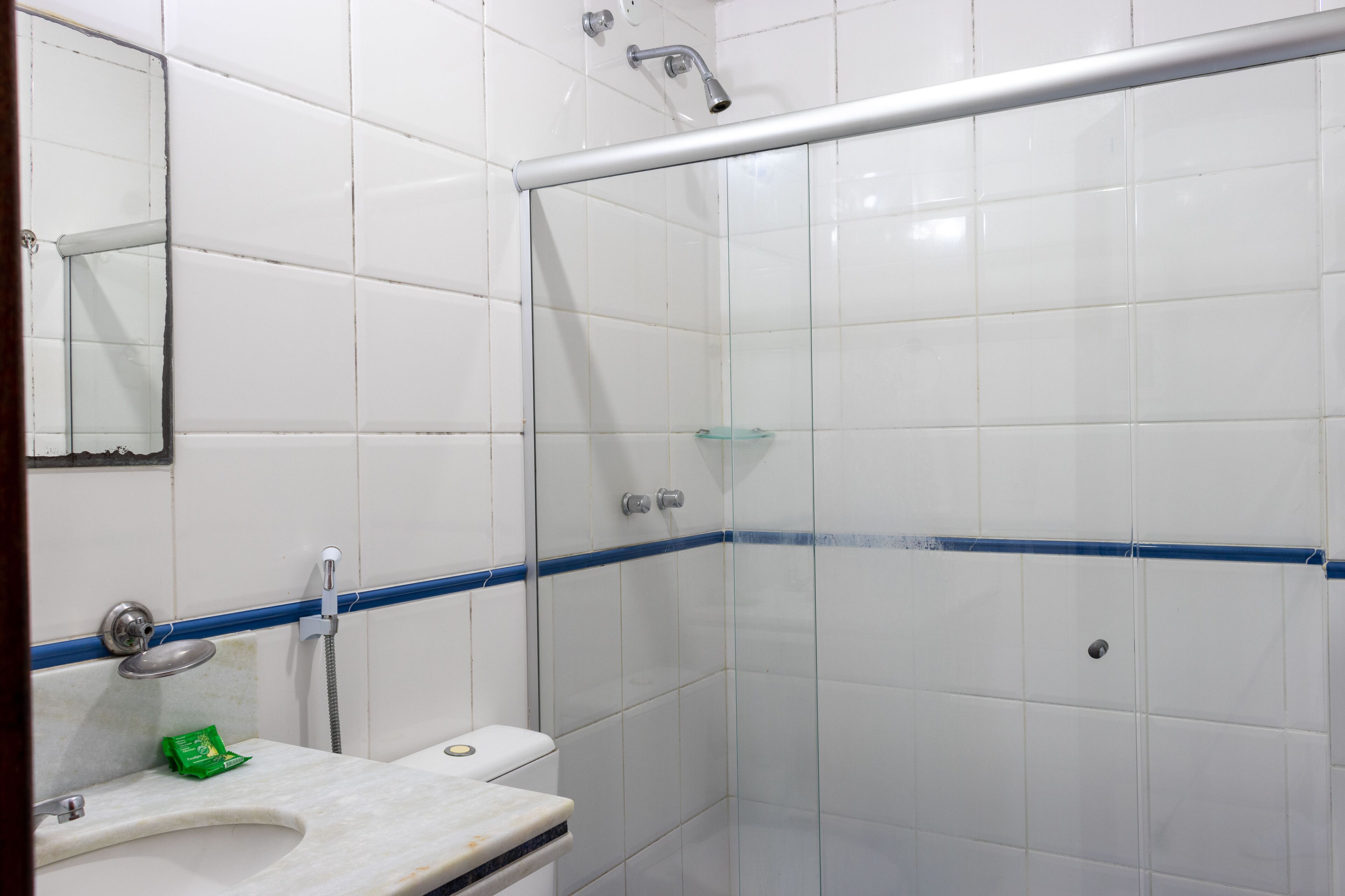 A bathroom with white tiled walls, a glass shower enclosure, a white sink with a green soap dispenser, and a mirror above the sink.