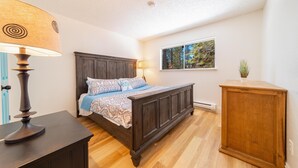 A bedroom with a wooden bed, a nightstand, a lamp, a window with a view of trees, and a small cabinet. - Luna Lodge - 4BR, 2BA Close to Hike, Bike (South Lake Tahoe)