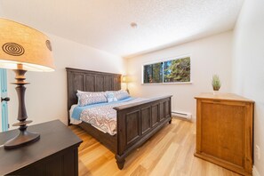 Une chambre à coucher avec un lit en bois, une table de chevet, une lampe, une fenêtre donnant sur des arbres et une petite armoire.