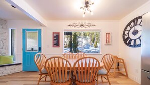 A dining area with a wooden table and chairs, a blue door, a large wall clock, and a window with a view of trees. - Luna Lodge - 4BR, 2BA Close to Hike, Bike (South Lake Tahoe)