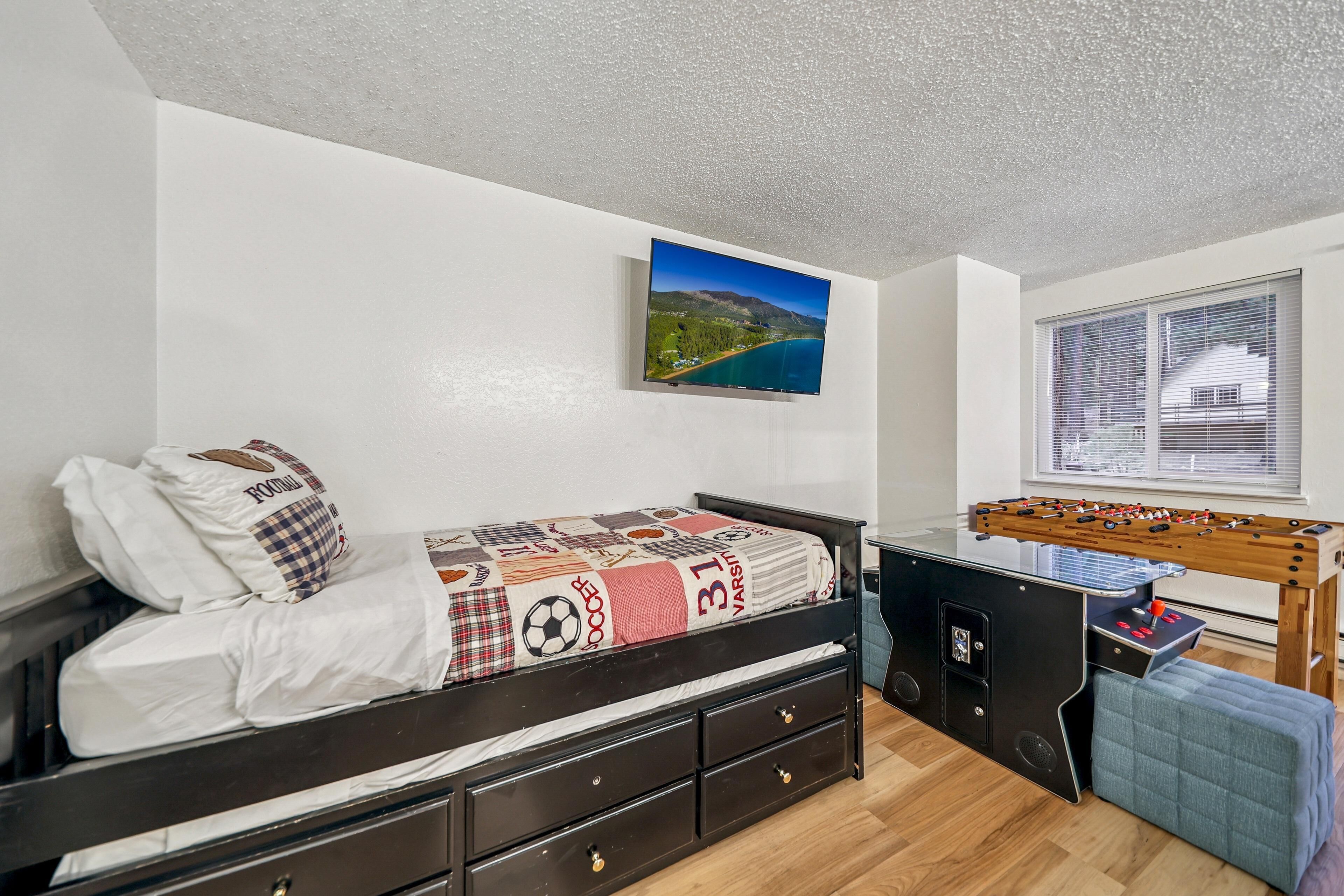 Un dormitorio con una cama, mesa de fútbol, ventana con persianas y un cuadro en la pared.