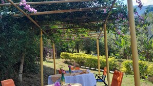 Een terras met tafels bedekt met beige tafelkleden, stoelen met oranje en blauwe kussens, en een pergola vol paarse bloemen.