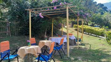 Et udendørs siddeområde med borde og stole, omgivet af grøn vegetation og en pergola.