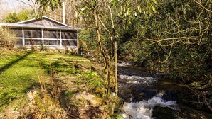 A house with a porch overlooking a flowing stream, surrounded by dense greenery. - "UNWIND BY THE WATER'S EDGE" Beautiful 2/1 Cabin w/ Hot Tub on Roaring Creek! (Bryson City)