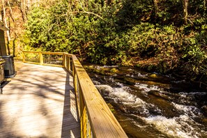A wooden deck overlooking a flowing river, surrounded by dense forest. - "UNWIND BY THE WATER'S EDGE" Beautiful 2/1 Cabin w/ Hot Tub on Roaring Creek! (Bryson City)