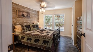 A bedroom with a wooden headboard, a quilted bedspread with bear designs, a ceiling fan, and a window with a view of trees. - "UNWIND BY THE WATER'S EDGE" Beautiful 2/1 Cabin w/ Hot Tub on Roaring Creek! (Bryson City)