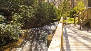 A wooden deck overlooking a stream with dense foliage on one side and a building with a staircase on the other. - "UNWIND BY THE WATER'S EDGE" Beautiful 2/1 Cabin w/ Hot Tub on Roaring Creek! (Bryson City)