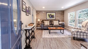 A living room with a leather sofa, a coffee table, and a plaid armchair. There is a fireplace with a mantel, a rug, and framed pictures on the wall. - "UNWIND BY THE WATER'S EDGE" Beautiful 2/1 Cabin w/ Hot Tub on Roaring Creek! (Bryson City)