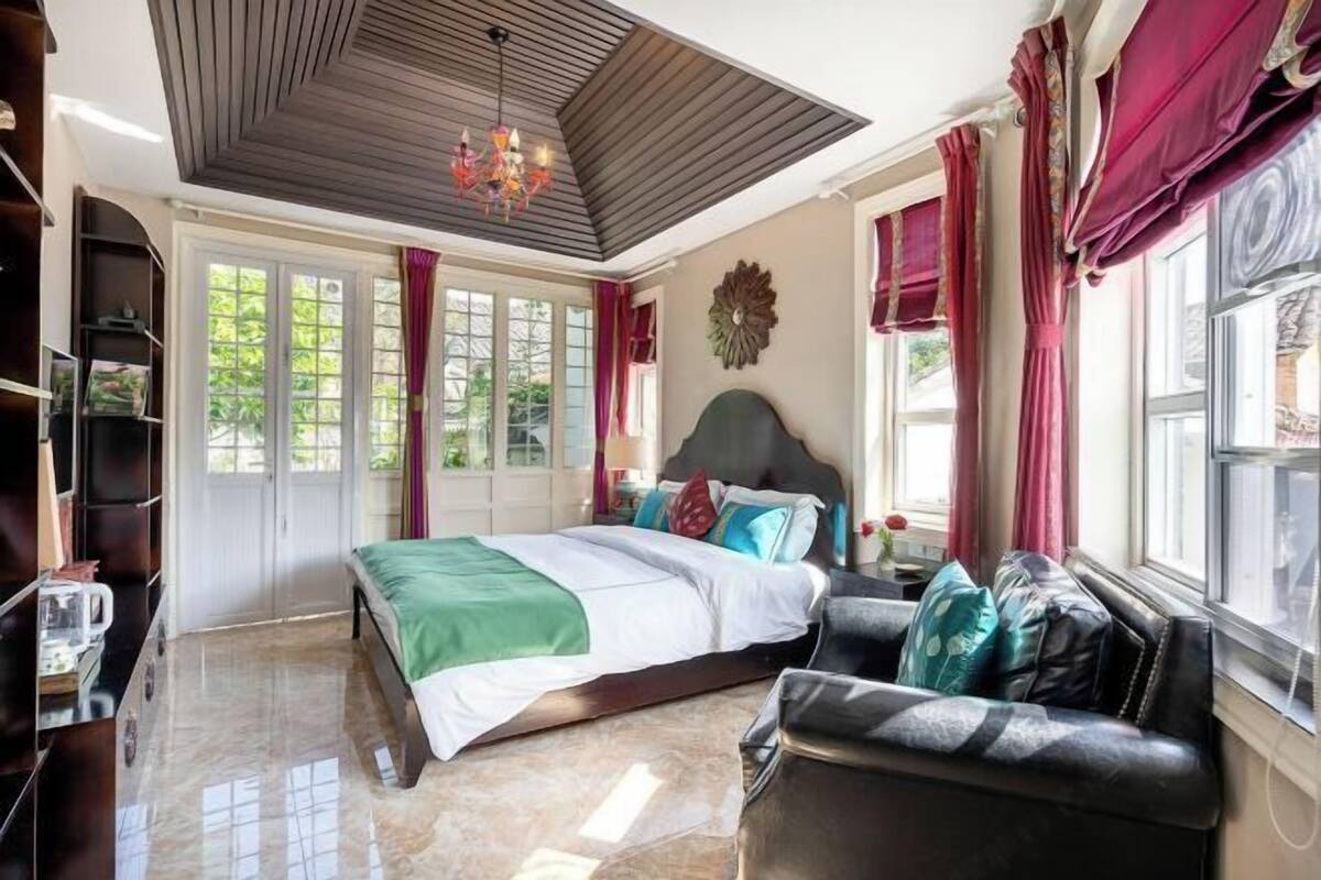 Un dormitorio con cama, sofá, candelabro y un ventanal grande con cortinas.