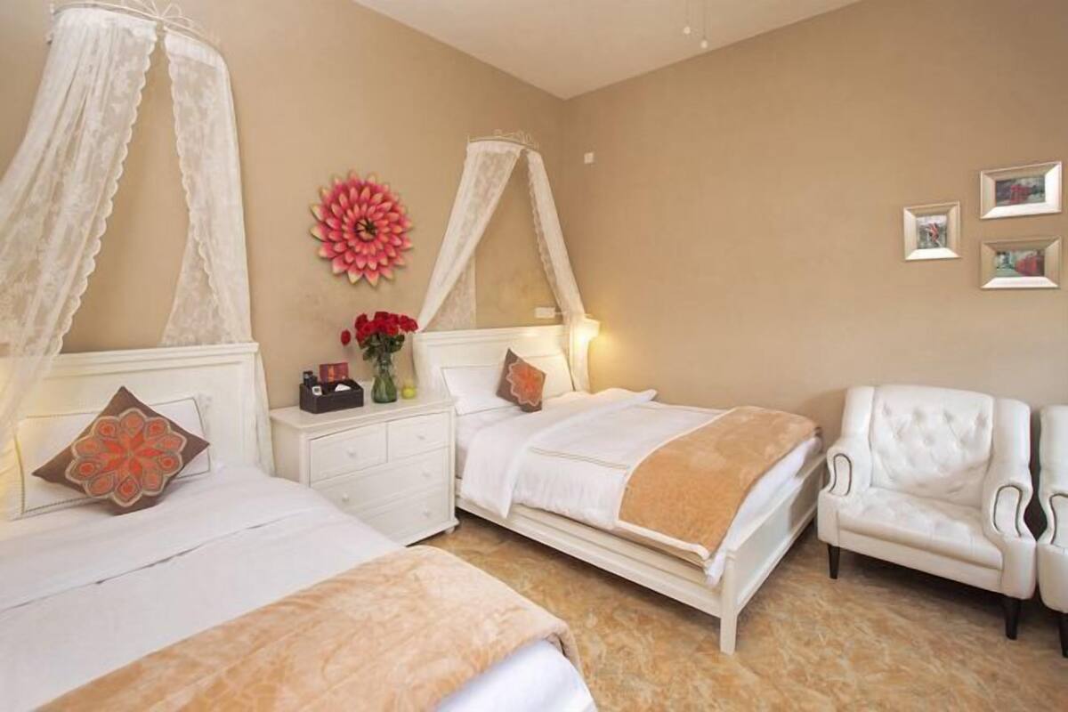Un dormitorio con dos camas, una silla blanca, una mesita de noche, una lámpara y un panel decorativo con flores.