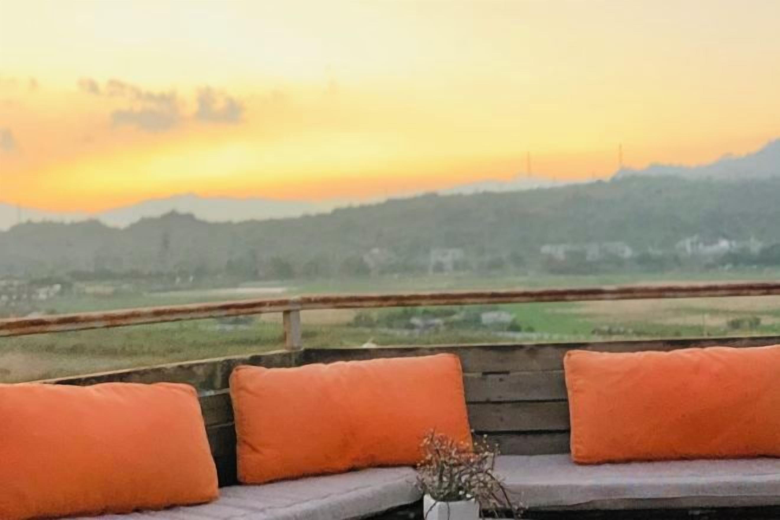 A bench with orange cushions and a vase with dried flowers, overlooking a landscape with a sunset.