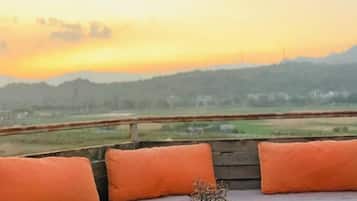 A bench with orange cushions and a vase with dried flowers, overlooking a landscape with a sunset.