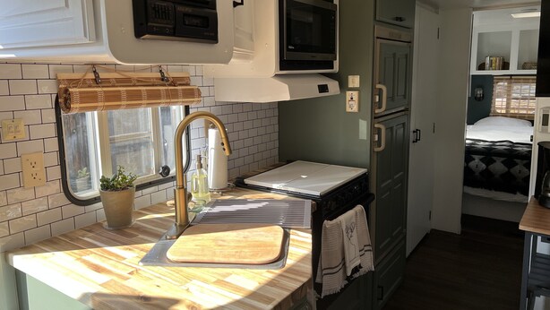 A compact kitchen with a sink, stove, microwave, and a small window with blinds. - Modern Vintage Camper w/ Peaceful Private Yard (Eugene)