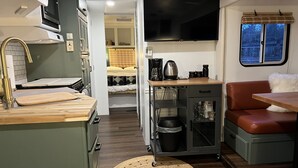 A compact kitchenette with a microwave, coffee maker, and storage cabinets. A small dining area with a table and a cushioned bench is visible. - Modern Vintage Camper w/ Peaceful Private Yard (Eugene)