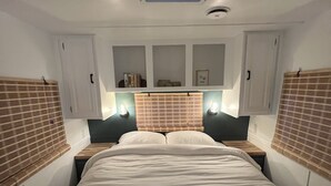 A compact, well-lit bedroom with a central bed, two bedside tables, a skylight, and built-in shelves with books and a framed picture. - Modern Vintage Camper w/ Peaceful Private Yard (Eugene)