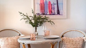 Un rincón comedor con sillas de mimbre, una mesa y un cuadro con tablas de surf y palmeras.