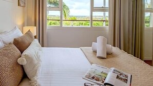 Un dormitorio con cama, almohadas, una lámpara de noche, una revista y una ventana con vistas a zonas verdes.
