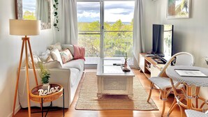 Un moderno salón con sofá, mesa de centro y sillas de mimbre. Hay un balcón con vistas a zonas verdes.