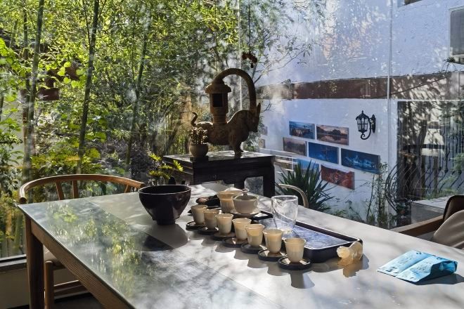 Une table dressée avec des théières, des tasses et une théière, placée sur une table en verre à l’extérieur, avec vue sur des arbres et un mur orné de tableaux.
