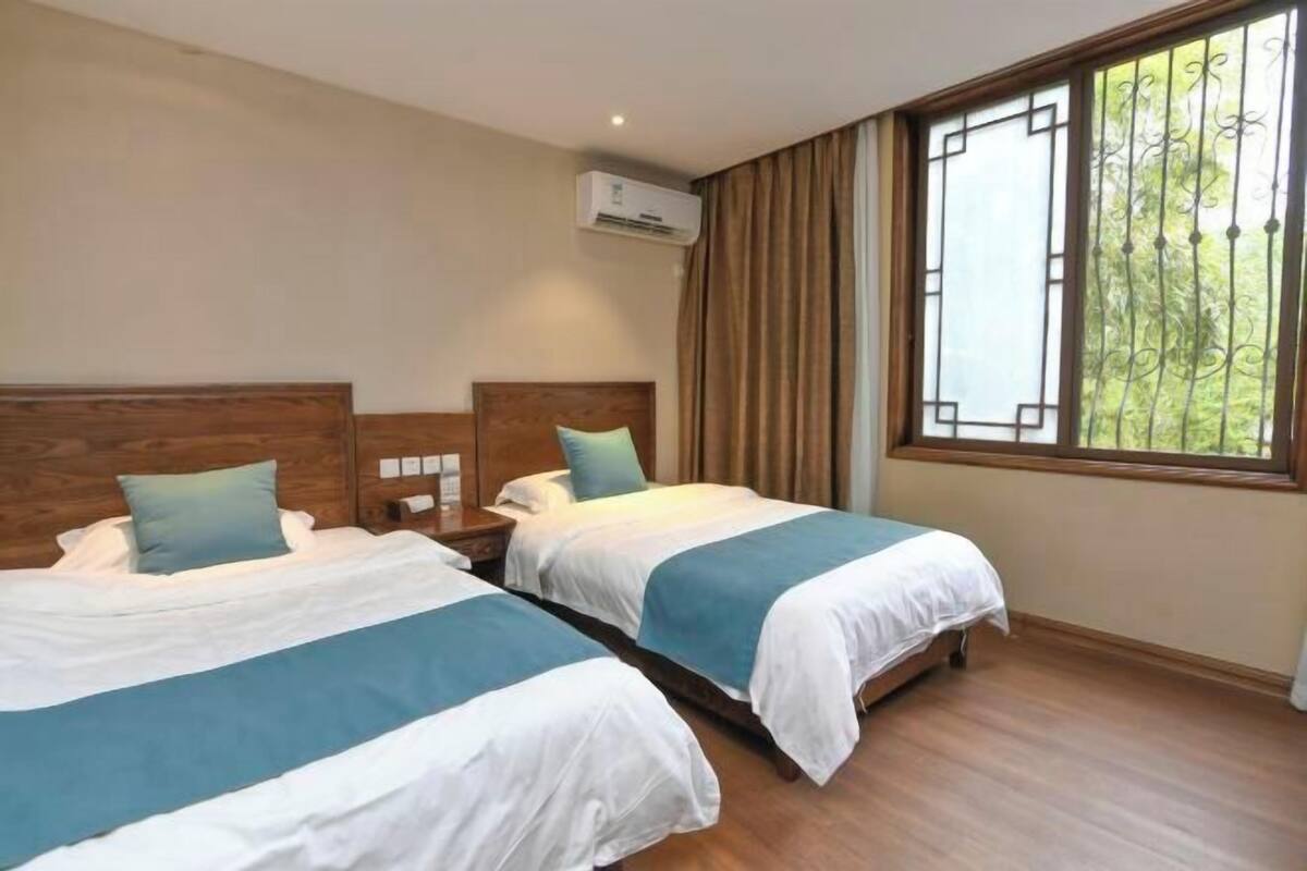 Habitación de hotel con dos camas, cabeceros de madera, un ventanal grande con cortinas y un equipo de aire acondicionado.
