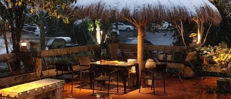 Un área de comedor al aire libre con un toldo de paja, asientos de piedra y suelo empedrado.