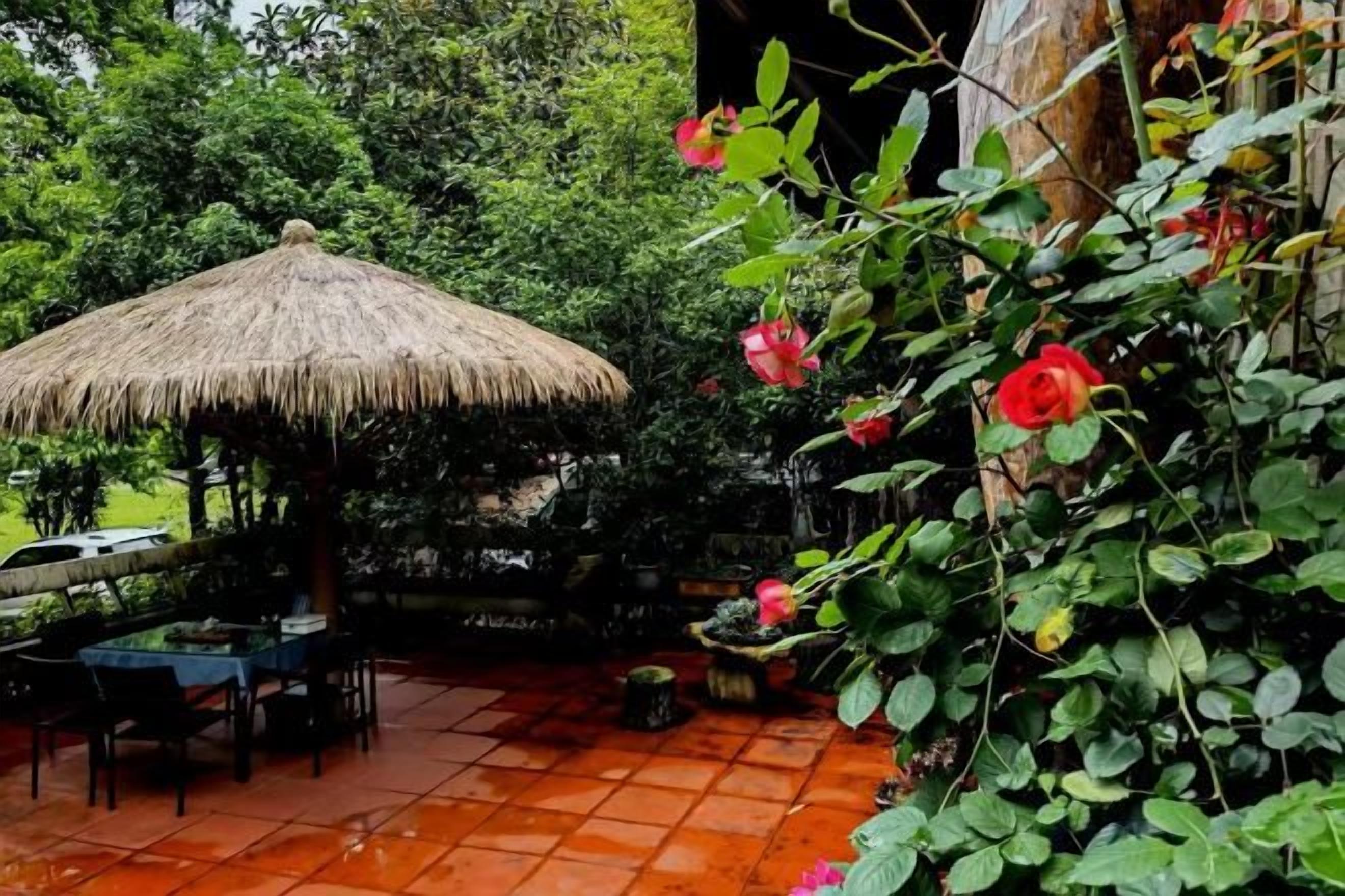 An outdoor dining area with a thatched roof, surrounded by lush greenery and a tiled floor.