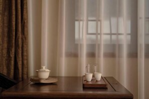 Una mesa con un hervidor, tazas y una bandeja en una habitación con cortinas translúcidas y una ventana.