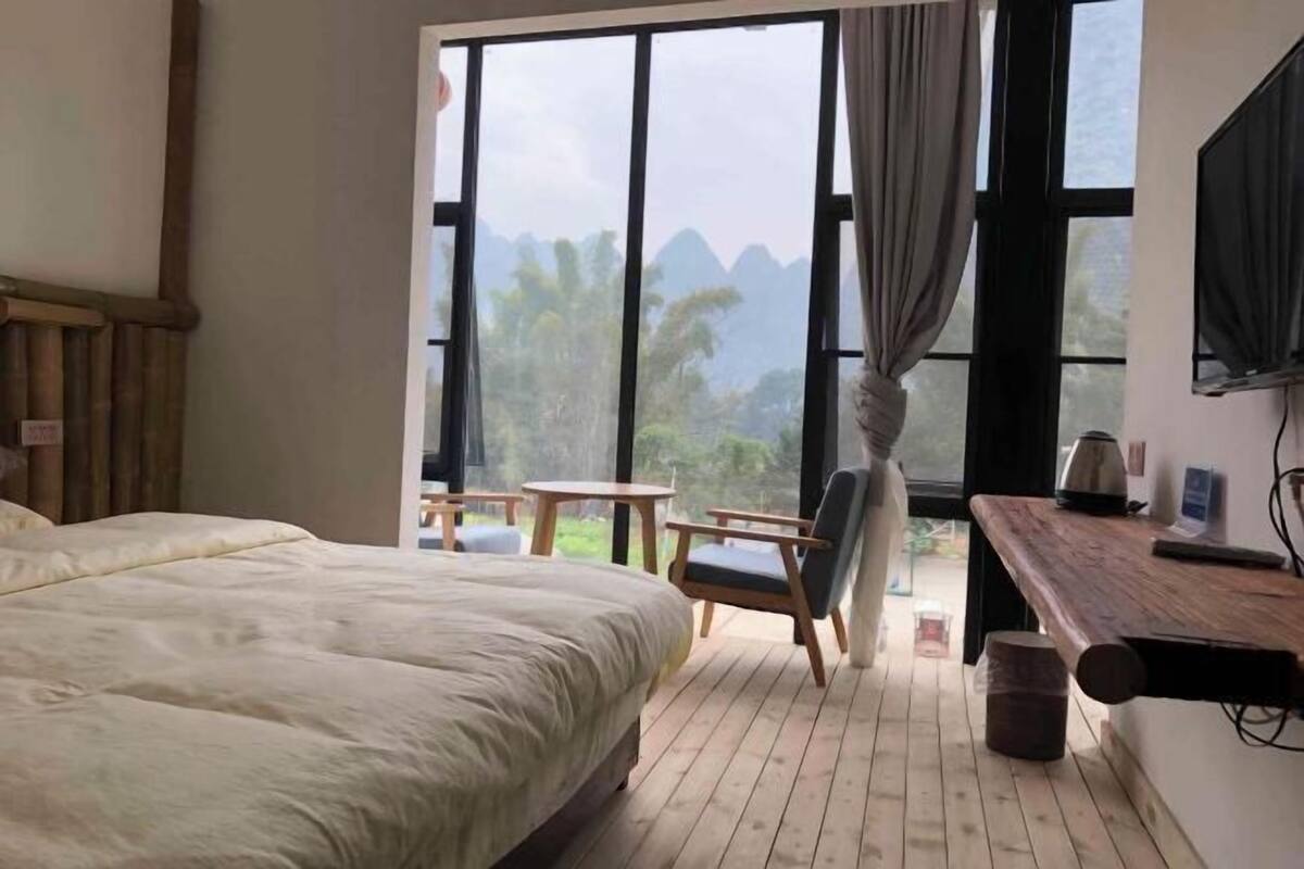 Quarto com cama grande, escrivaninha de madeira, cadeira e vista para montanhas através de grandes janelas.
