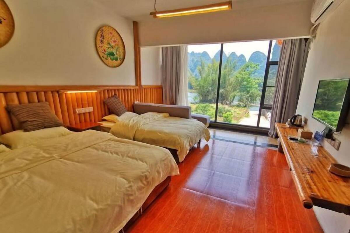 Quarto de hotel com duas camas, uma secretária de madeira e uma janela grande com vista para áreas verdes e montanhas.