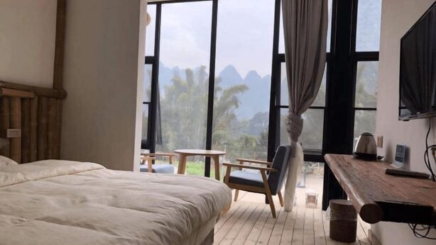 A bedroom with a large bed, a wooden desk, a chair, and a view of mountains through large windows. - Laozheng Homestay (Yangshuo)