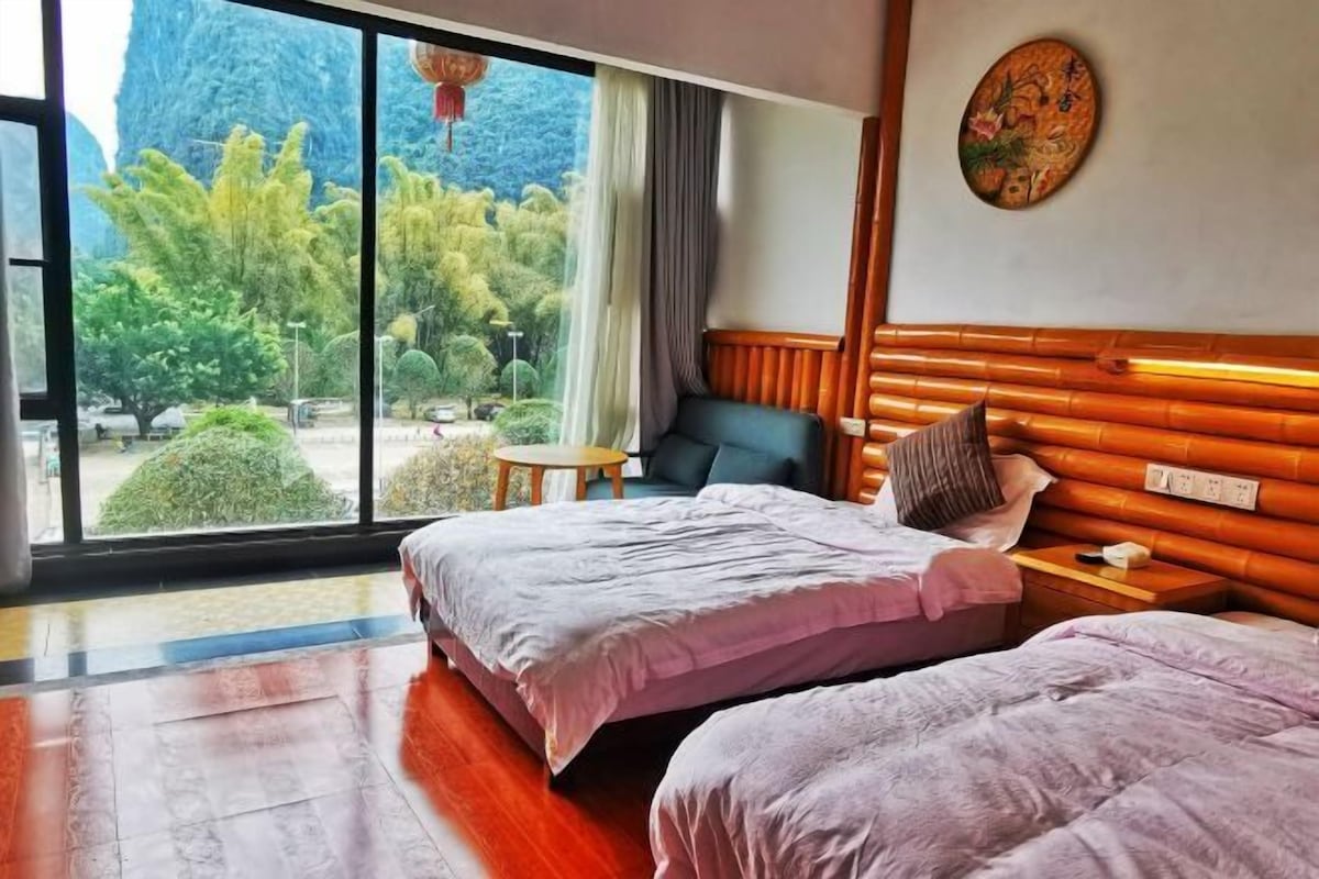 A hotel room with two beds, a view of greenery and mountains, a wooden headboard, and a small table.