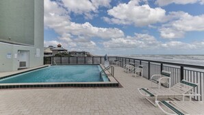 A rooftop pool with a view of the ocean, surrounded by lounge chairs and a building with a distinctive roof.