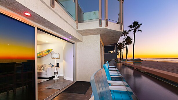 A modern hotel room with a balcony overlooking the beach, a poolside seating area, and a surfboard display. - Luxury Oceanfront, Two-Level Condo with Stunning View, Decks, Fast WiFi & Fireplace! (San Diego)