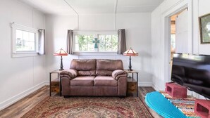 Una sala de estar con un sofá de cuero marrón, dos lámparas, un televisor sobre un mueble bajo y una alfombra estampada.