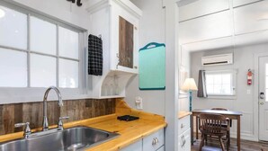 Una cocina con encimera de madera, pileta de acero inoxidable y armarios blancos. Se observa una ventana con vistas, una mesa con sillas y un equipo de aire acondicionado en la pared.