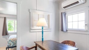 Una habitación con una mesa y sillas de madera, una lámpara, una cama con ropa de cama estampada y un equipo de aire acondicionado.