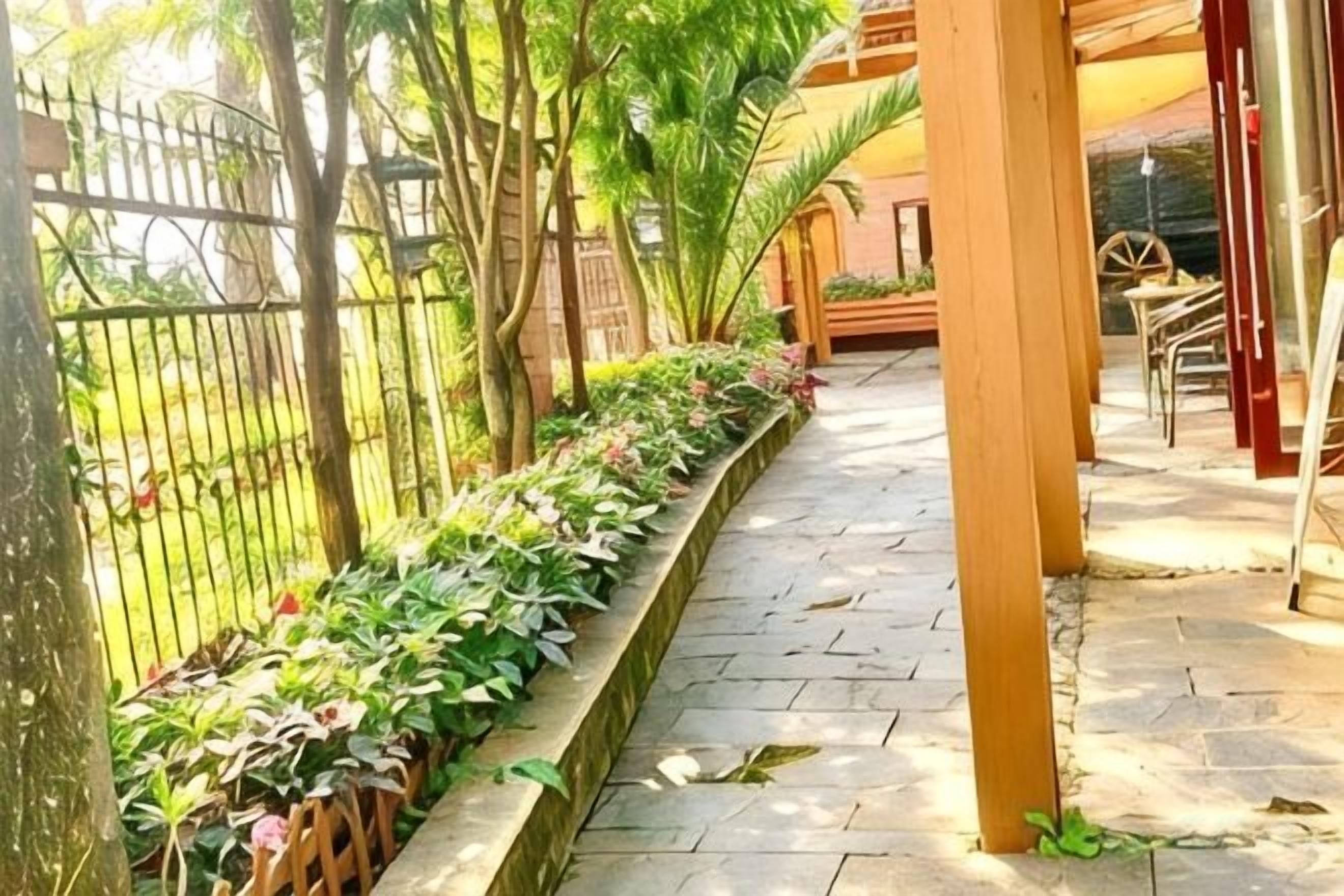 Ein gepflasterter Weg mit einem Grünpflanzen-Blumenkasten, einer hölzernen Pergola und einem von Bäumen gesäumten, eingezäunten Bereich.