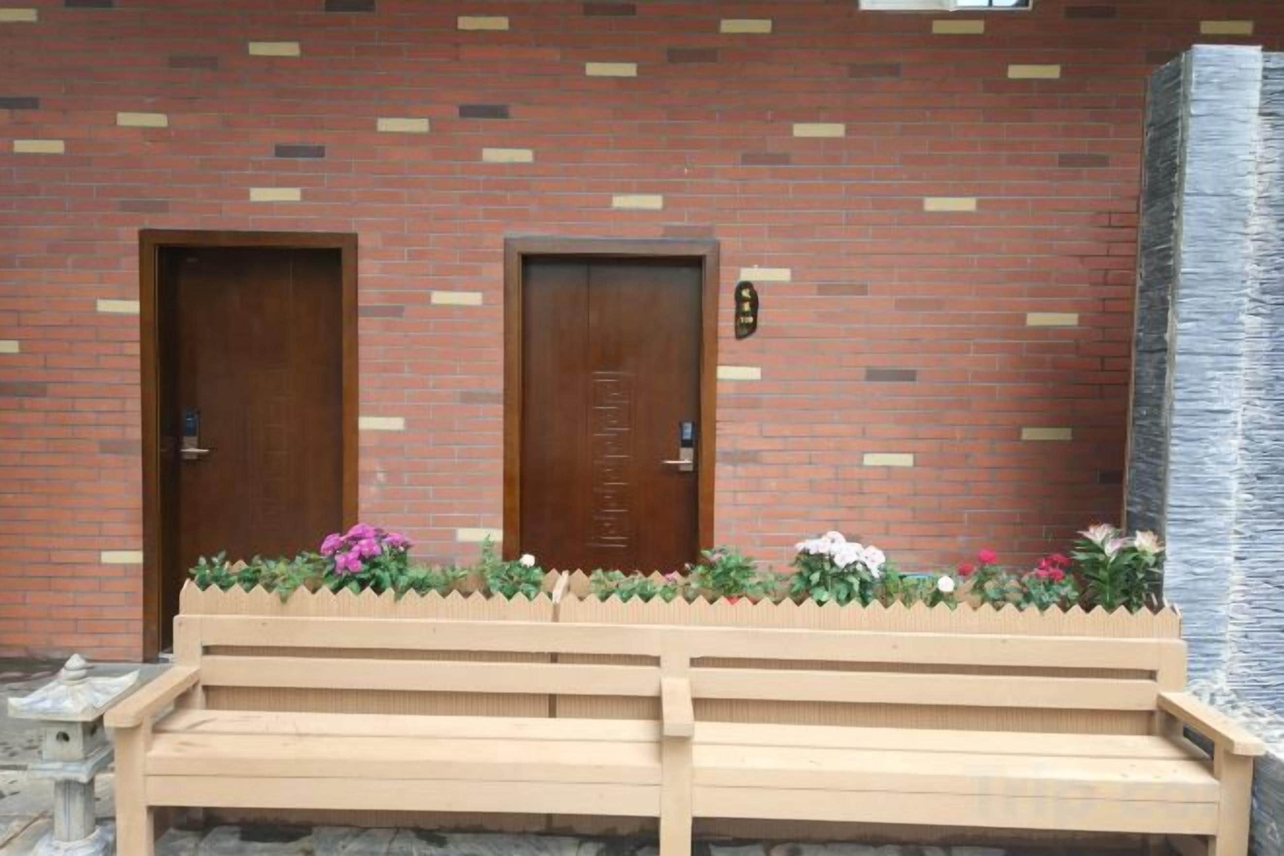 A building with two wooden doors, a bench, and a flower bed with pink flowers.