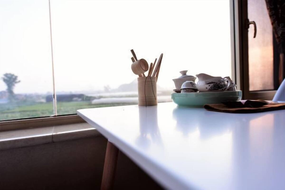 A table with a bowl of chopsticks and teapots, a window with a view of a green landscape.