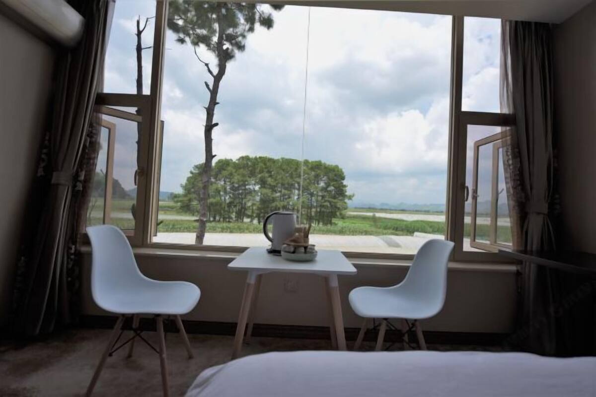 A hotel room with a bed, two white chairs, a small table with a kettle and a cup, and a large window with a view of trees and a cloudy sky.