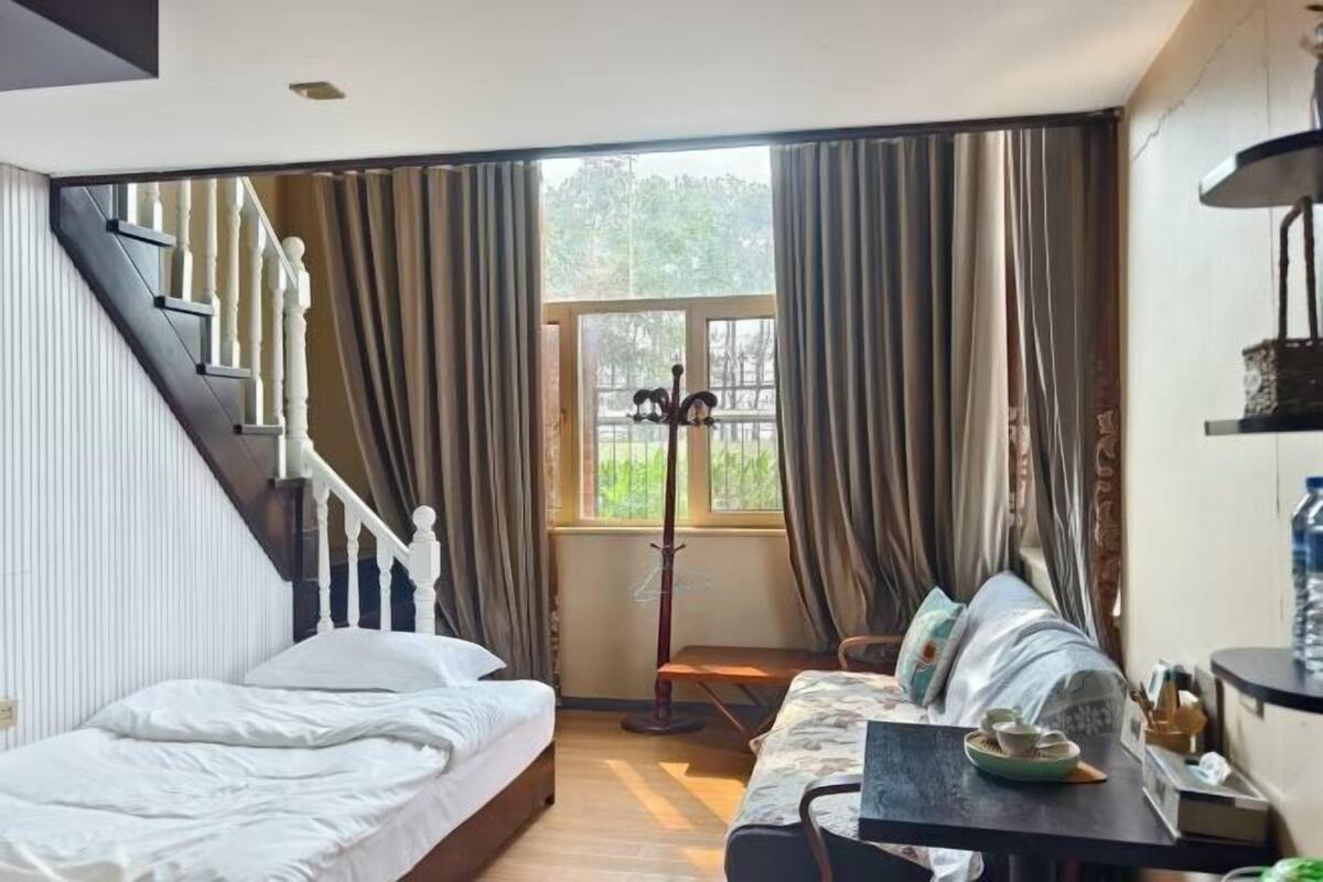A bedroom with a bed, a sofa, a table, a window with curtains, a coat rack, and a staircase.