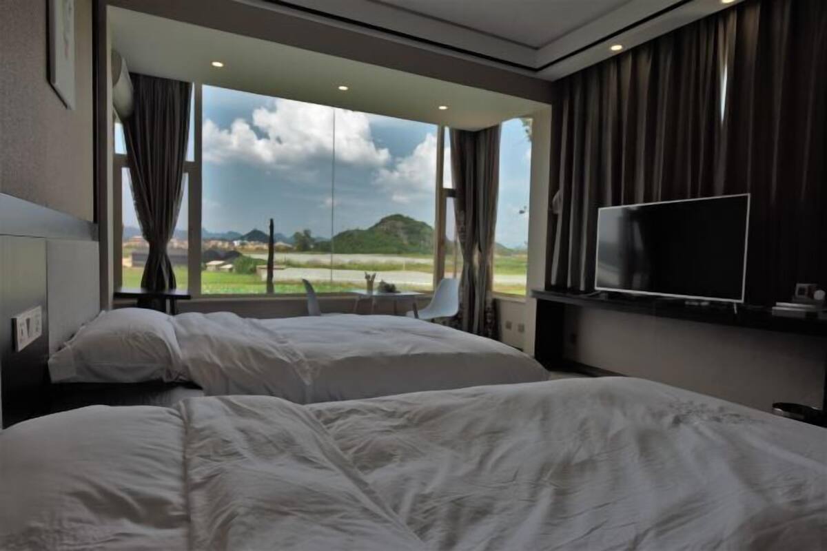 A hotel room with two beds, a TV, and a large window with a view of the countryside.