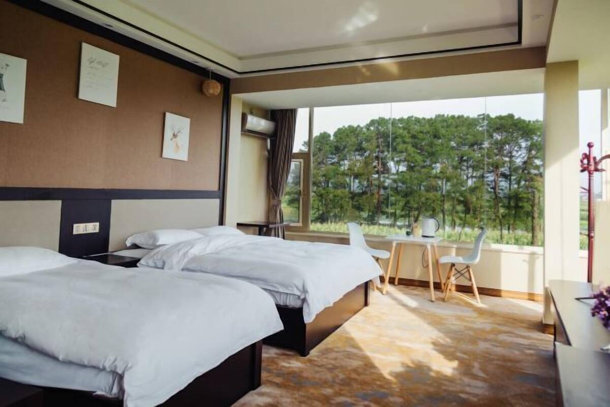 A hotel room with two beds, a desk, and a view of trees through large windows.