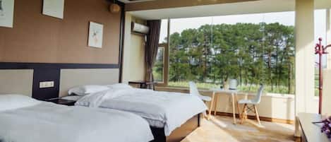 A hotel room with two beds, a desk, and a view of trees through large windows.
