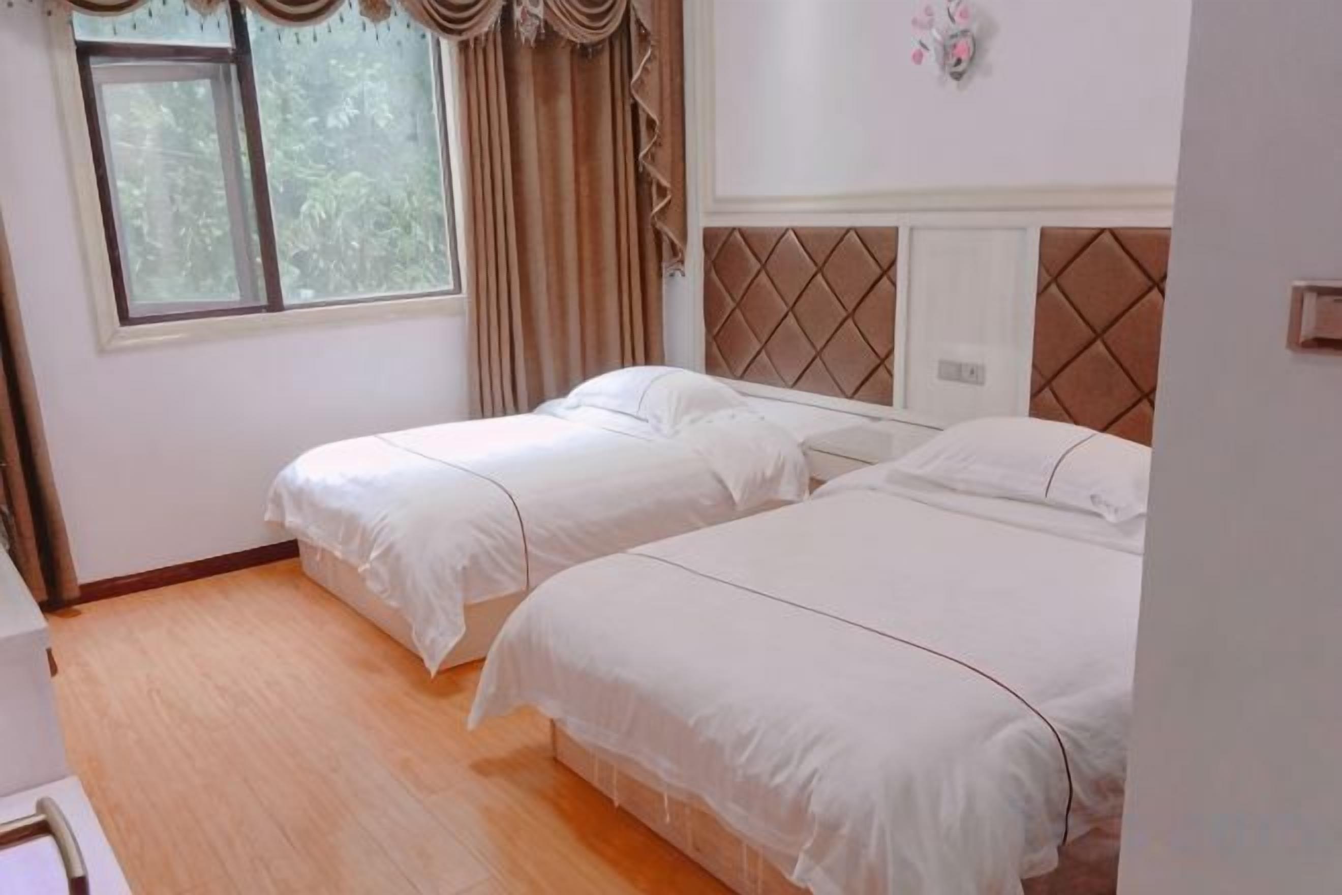 A room with two single beds, a window with curtains, a wooden floor, and a wall-mounted light switch.